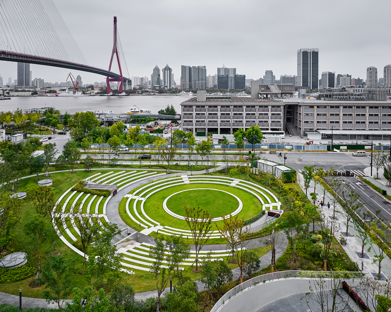 Concentric white-striped amphitheatre bowl surrounded by trees and river with bridge beyond under overcast sky
