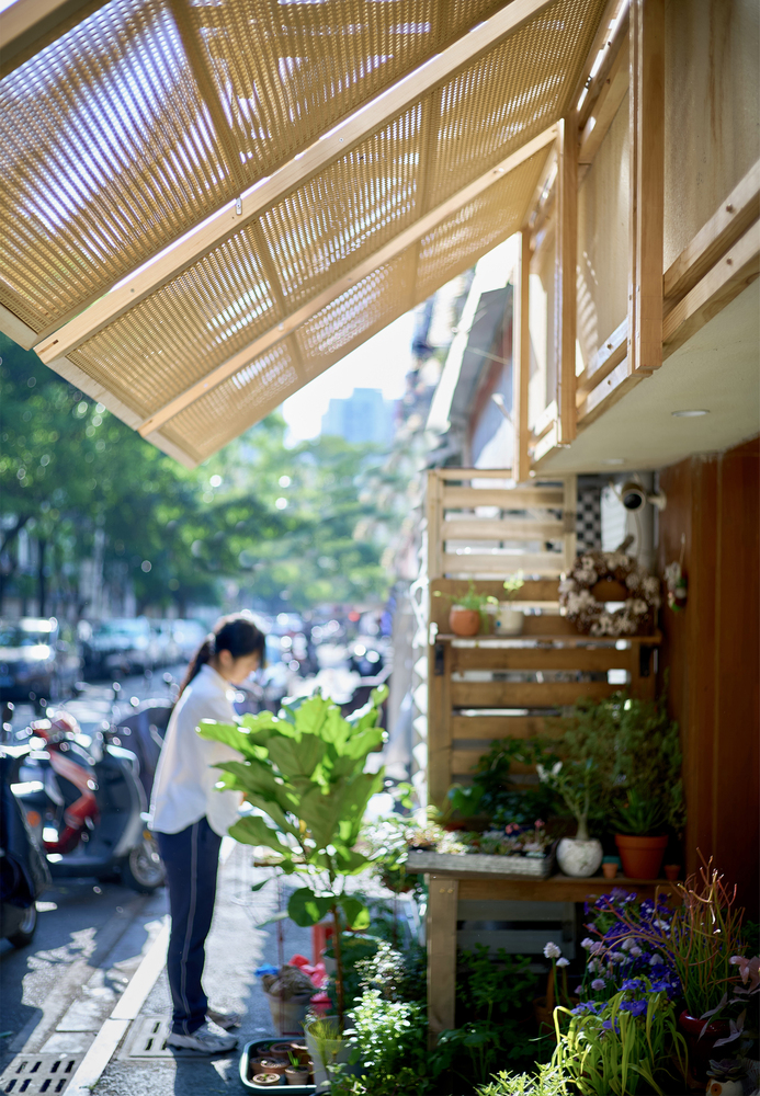 Person tending plants beneath the louvered timber canopy filtering dappled sunlight onto the sidewalk shop