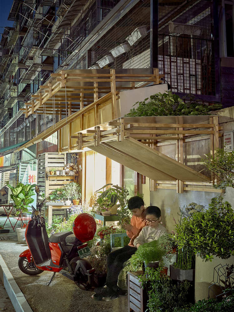 Evening view of the illuminated storefront with two people sitting among potted plants under the timber structure