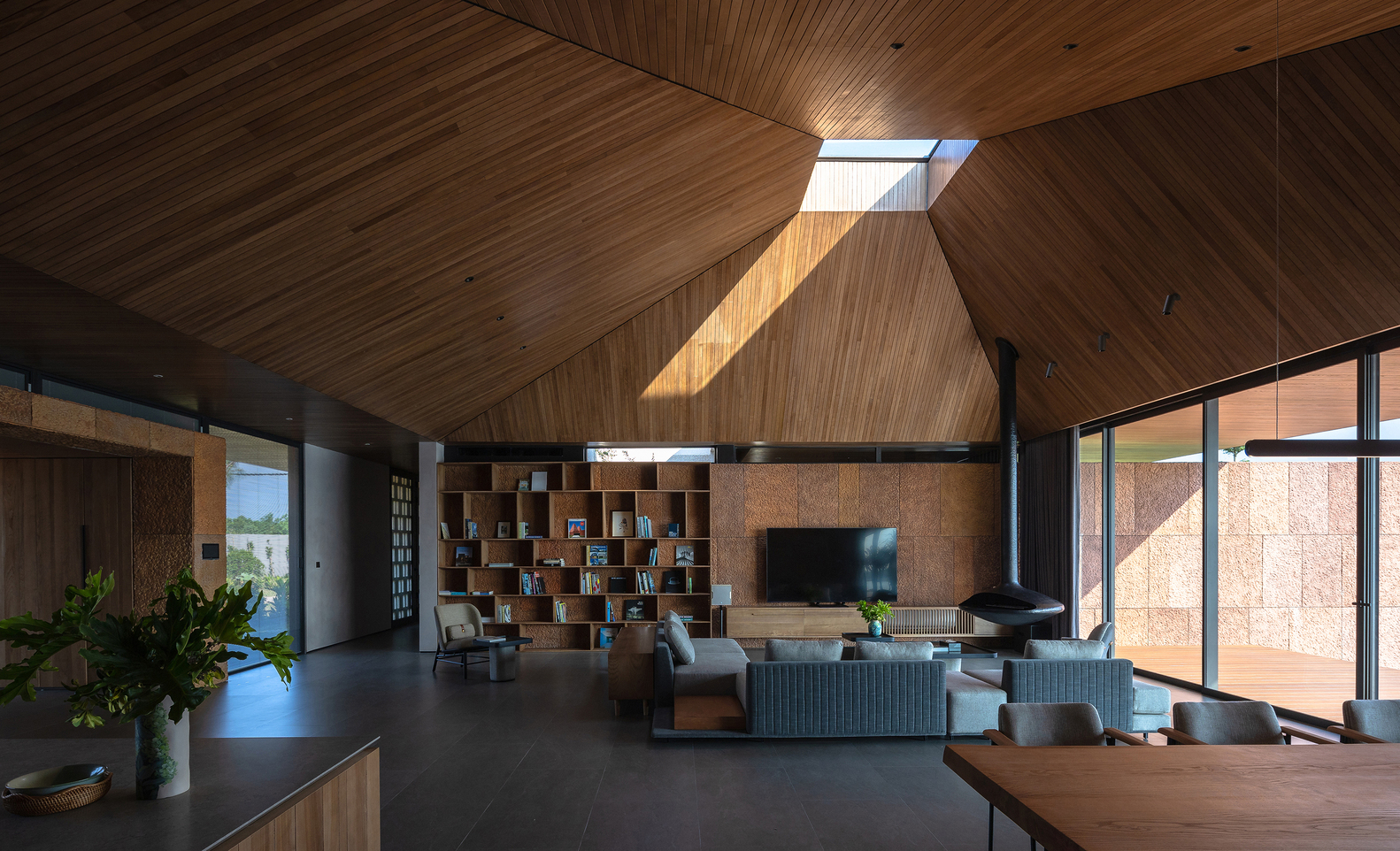 Open-plan living space with timber-clad vaulted ceiling and central skylight casting afternoon sunlight onto the modular shelving wall