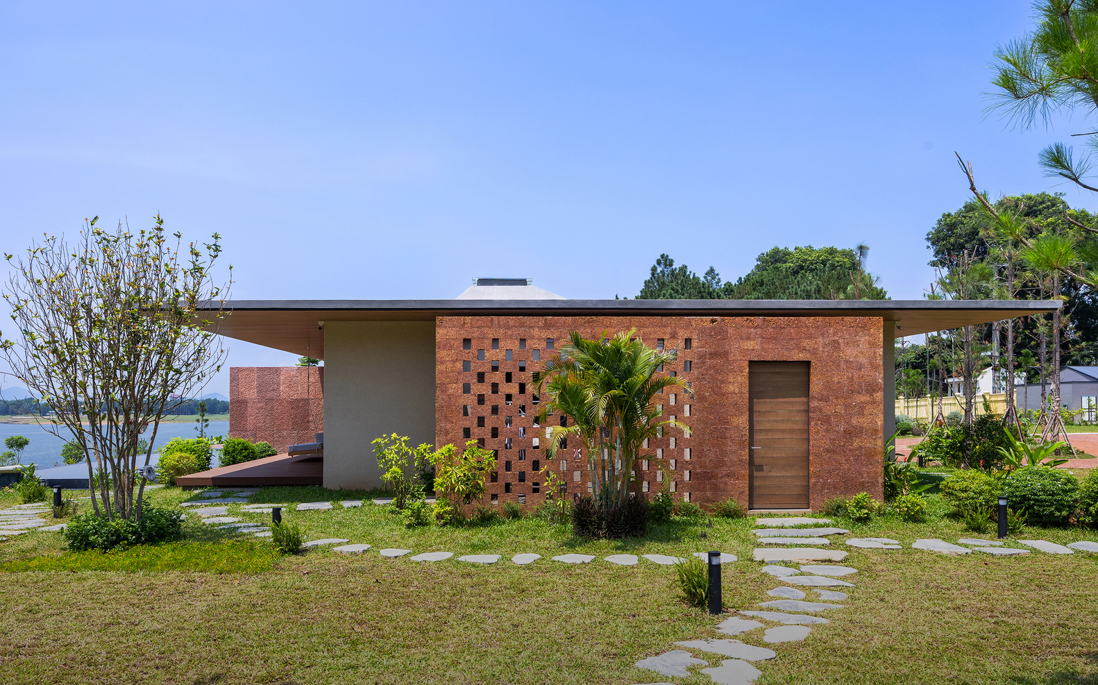 Red brick facade with perforated pattern and timber door behind stepping stones and planted shrubs in full sun