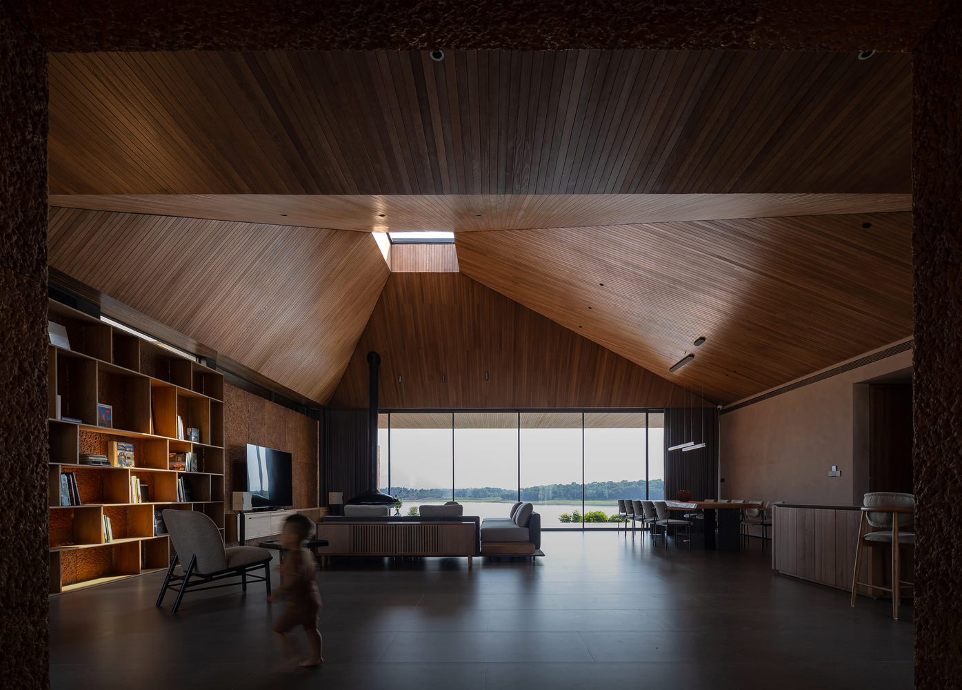Living room with faceted timber ceiling, clerestory window, and a child walking across the polished floor toward lake views