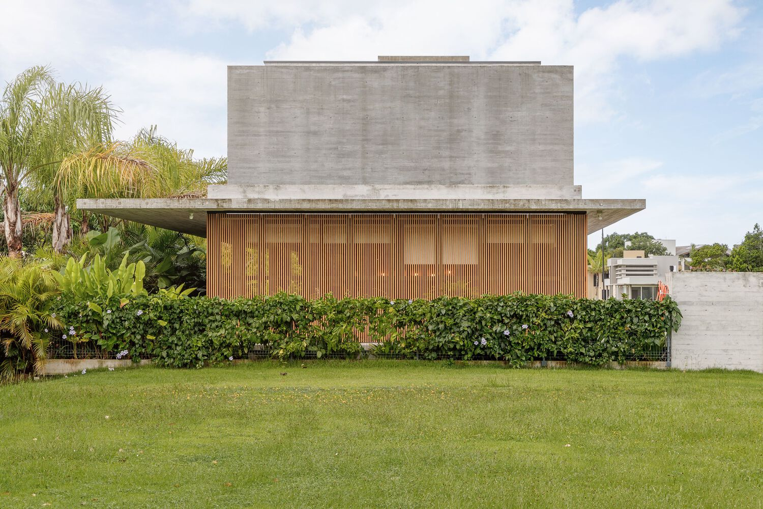 Stacked concrete volume above timber-clad lower level with landscaped hedge and palm trees