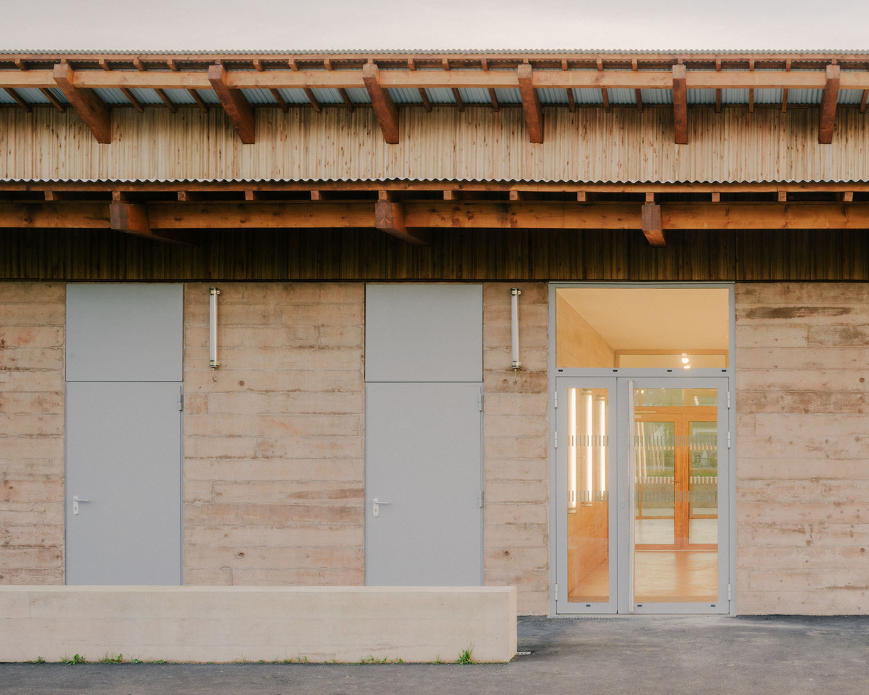 Entrance facade showing rammed earth walls with metal doors and glazed opening below exposed timber eaves