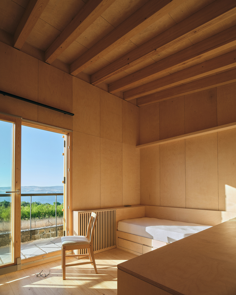 Interior bedroom with exposed timber beams and built-in plywood platform bed overlooking a vineyard and water