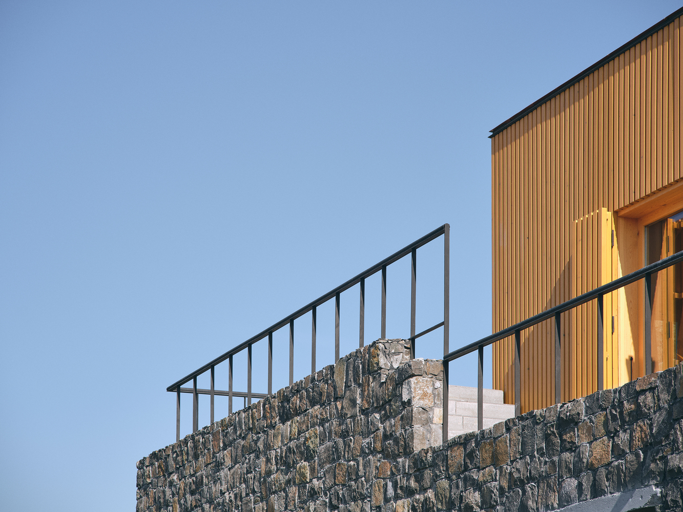 Detail of the steel railing atop a dark stone wall with vertical timber cladding beyond