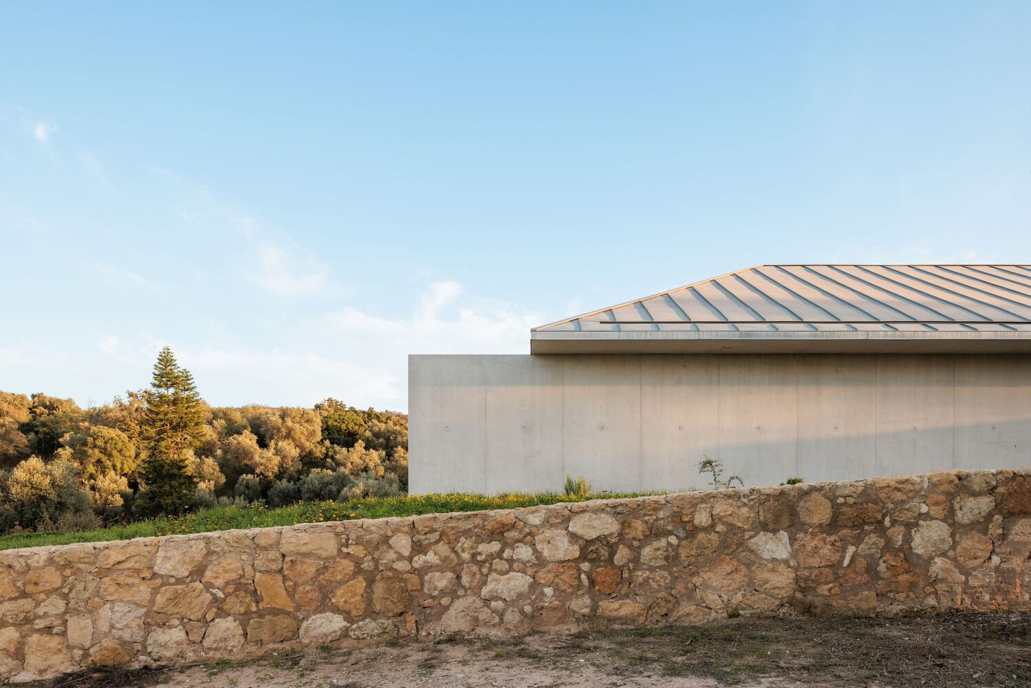 Metal standing seam roof edge above concrete wall with stone retaining wall in afternoon light