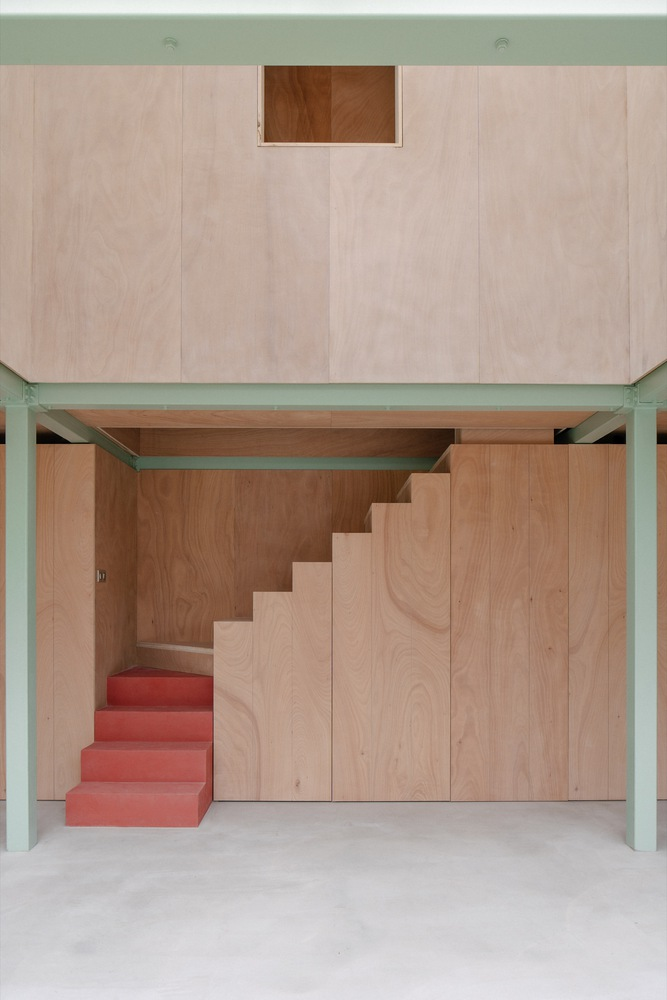 Plywood staircase with coral-painted treads tucked beneath a suspended box volume with green steel frame
