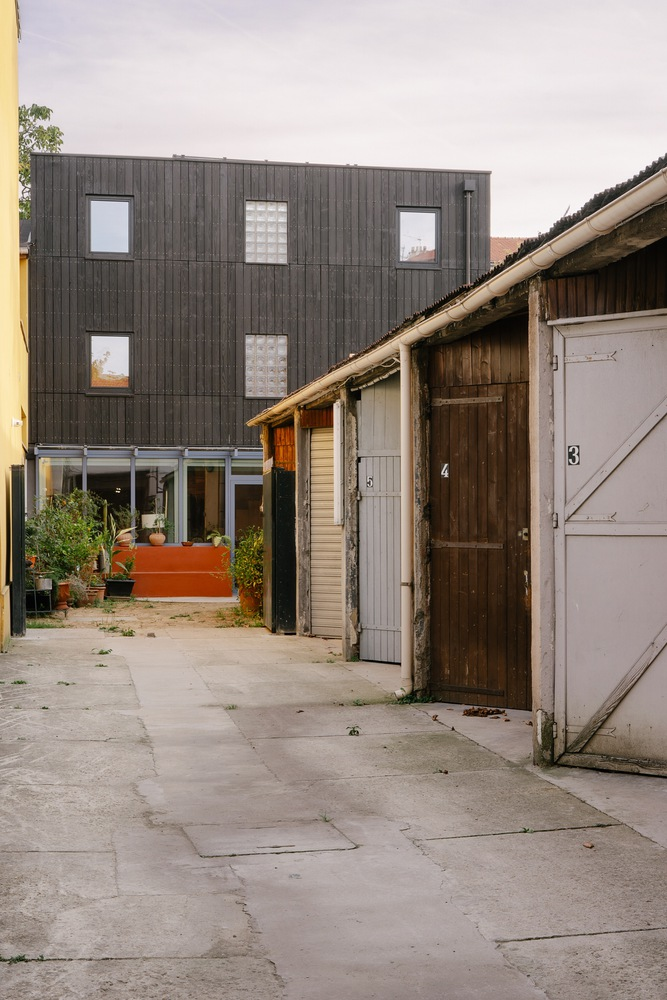 Dark timber-clad facade rising behind existing garage structures in a courtyard at dusk