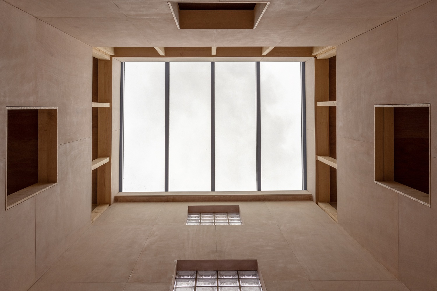 Looking up at a plywood-clad shaft with tall glazing and recessed timber niches on both sides