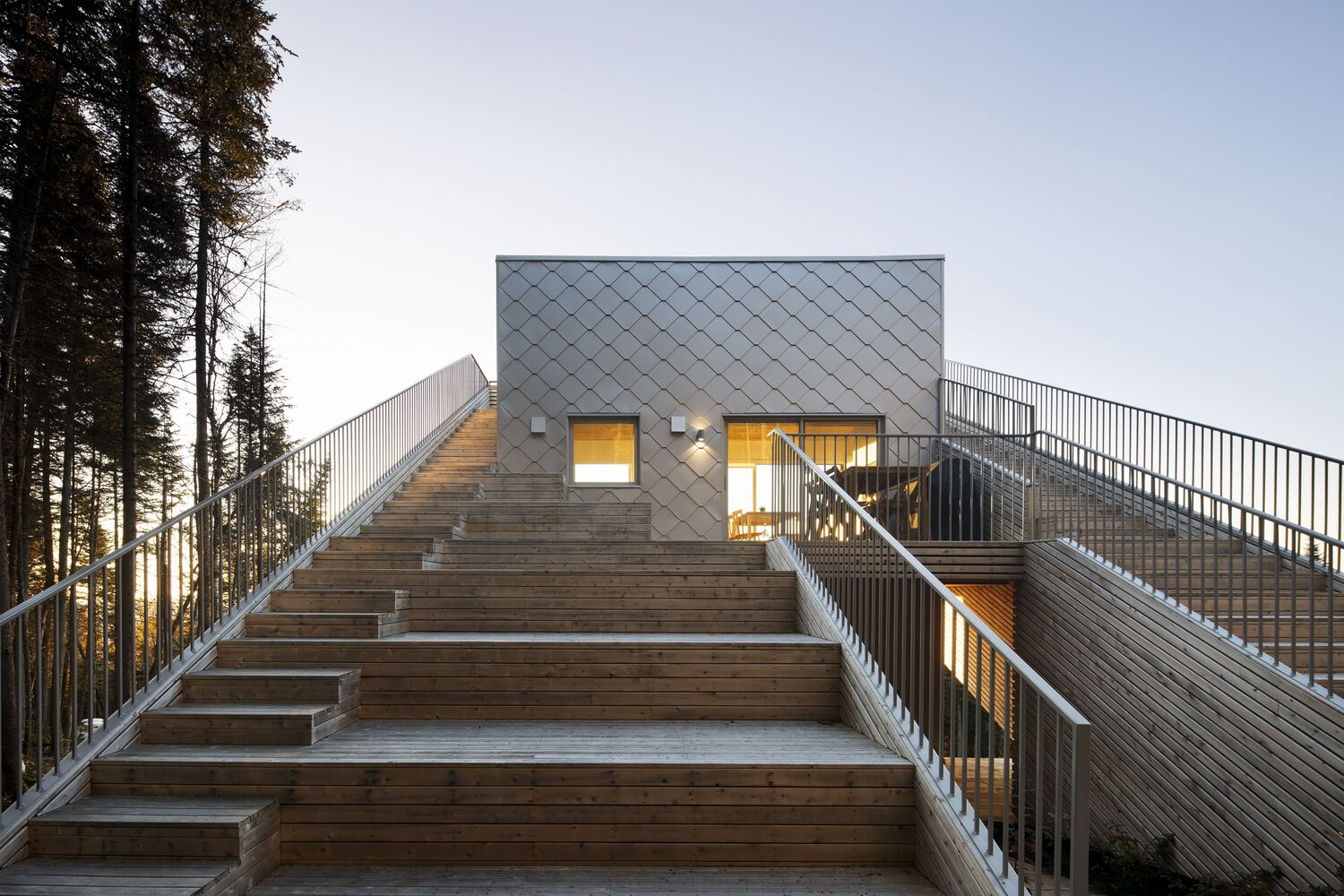 Timber staircase leading to upper volume clad in diamond-pattern metal panels at dusk