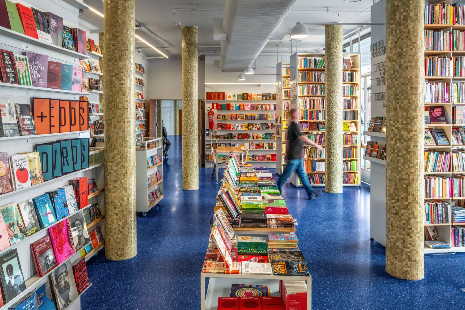 Bookstore interior with cork-clad columns, blue terrazzo flooring, and a figure browsing shelves