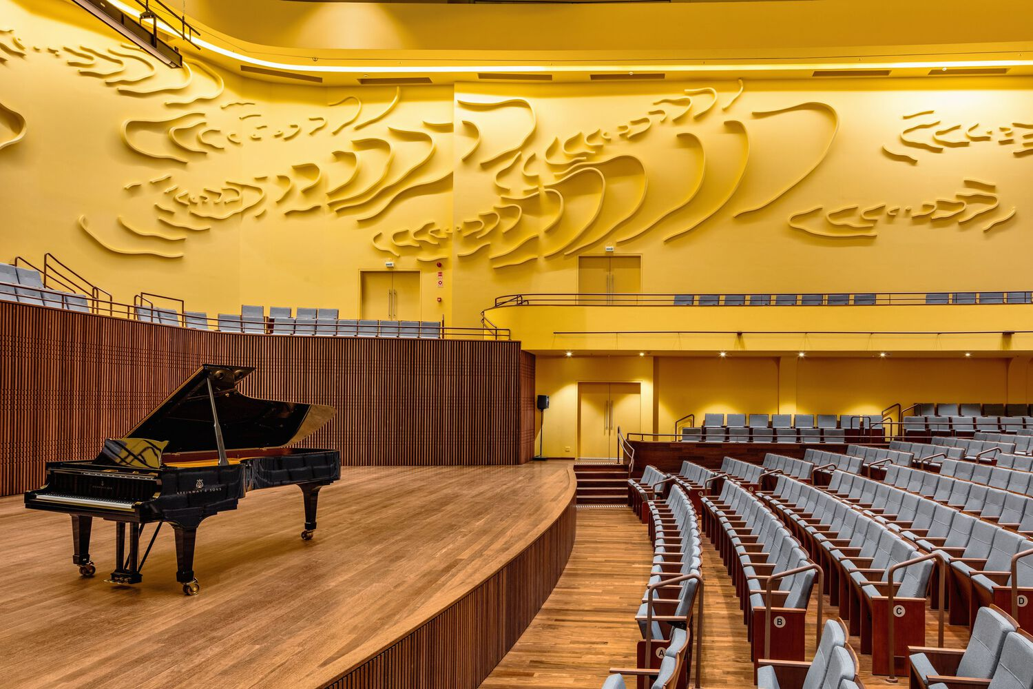 Grand piano on curved timber stage beneath the sculpted yellow wall and auditorium seating under warm lighting