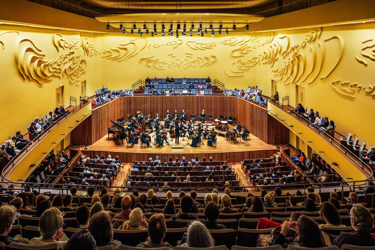 View from rear seating toward orchestra on stage framed by curved timber balcony and relief wall