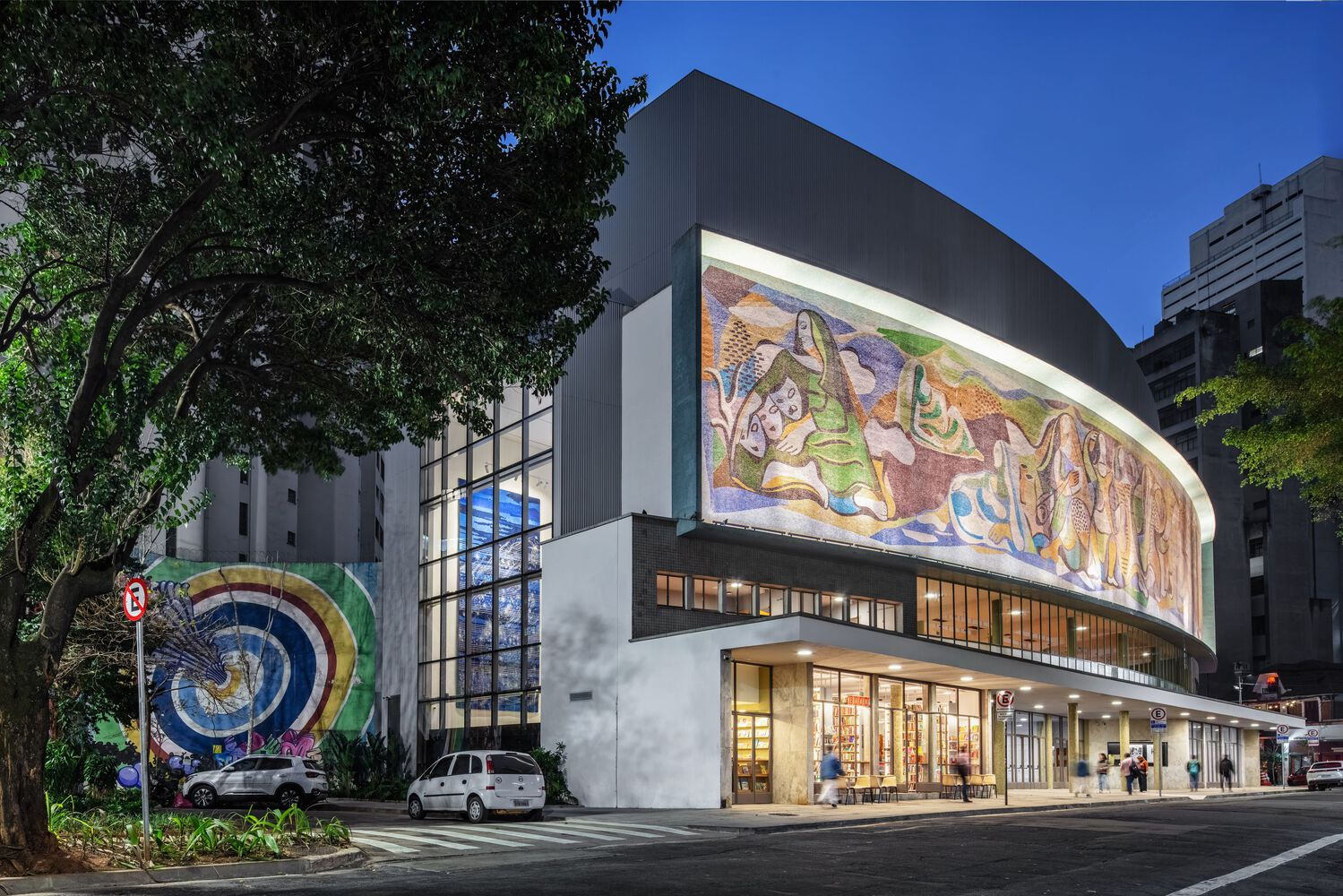 Corner view of the curved facade with mosaic mural illuminated at twilight beneath a blue sky