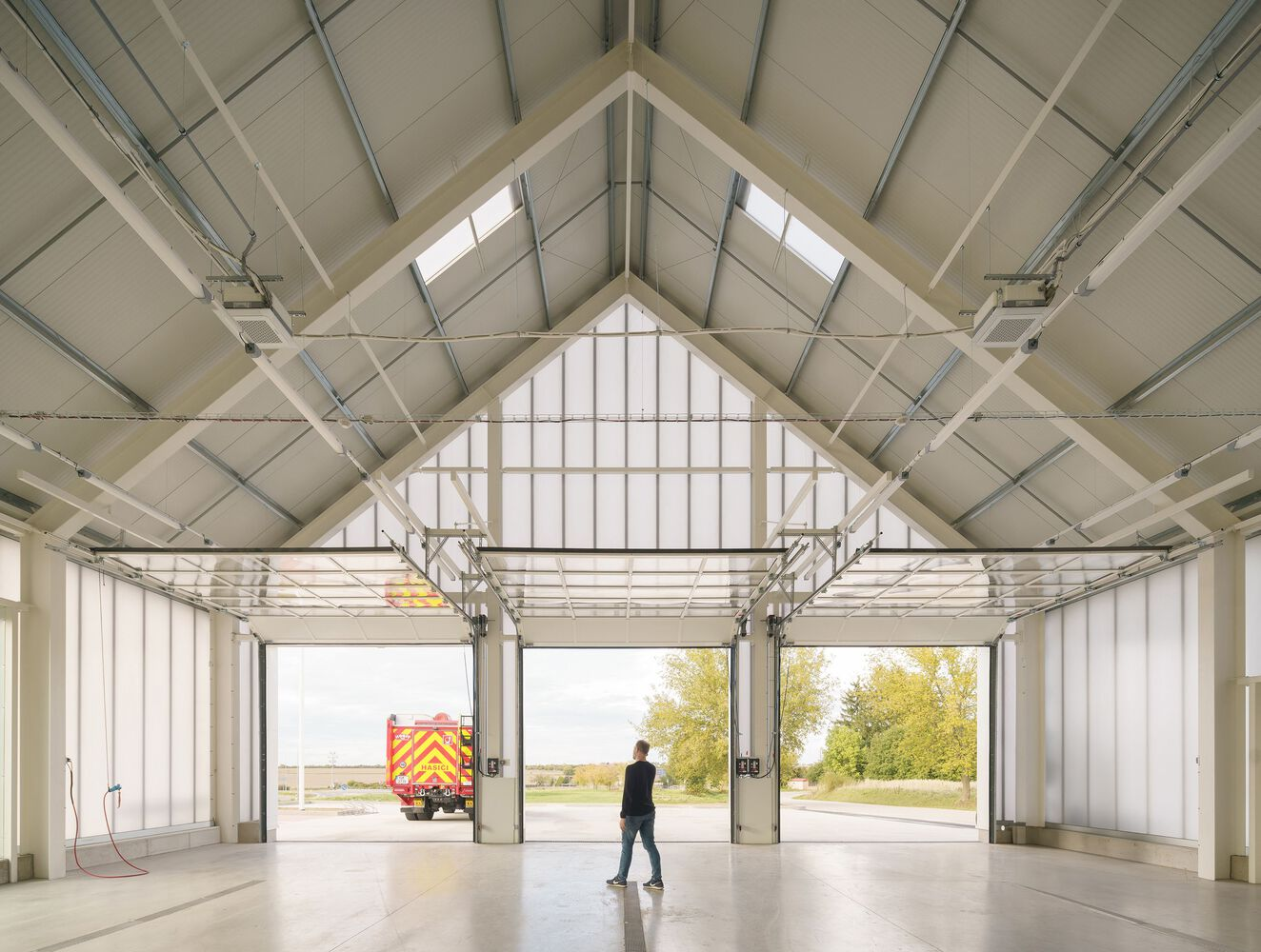 Interior of a fire station garage bay with exposed gabled roof structure and person walking toward open doors