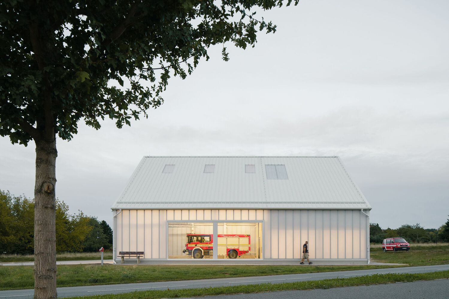 White corrugated metal building with illuminated translucent facade housing fire trucks at dusk