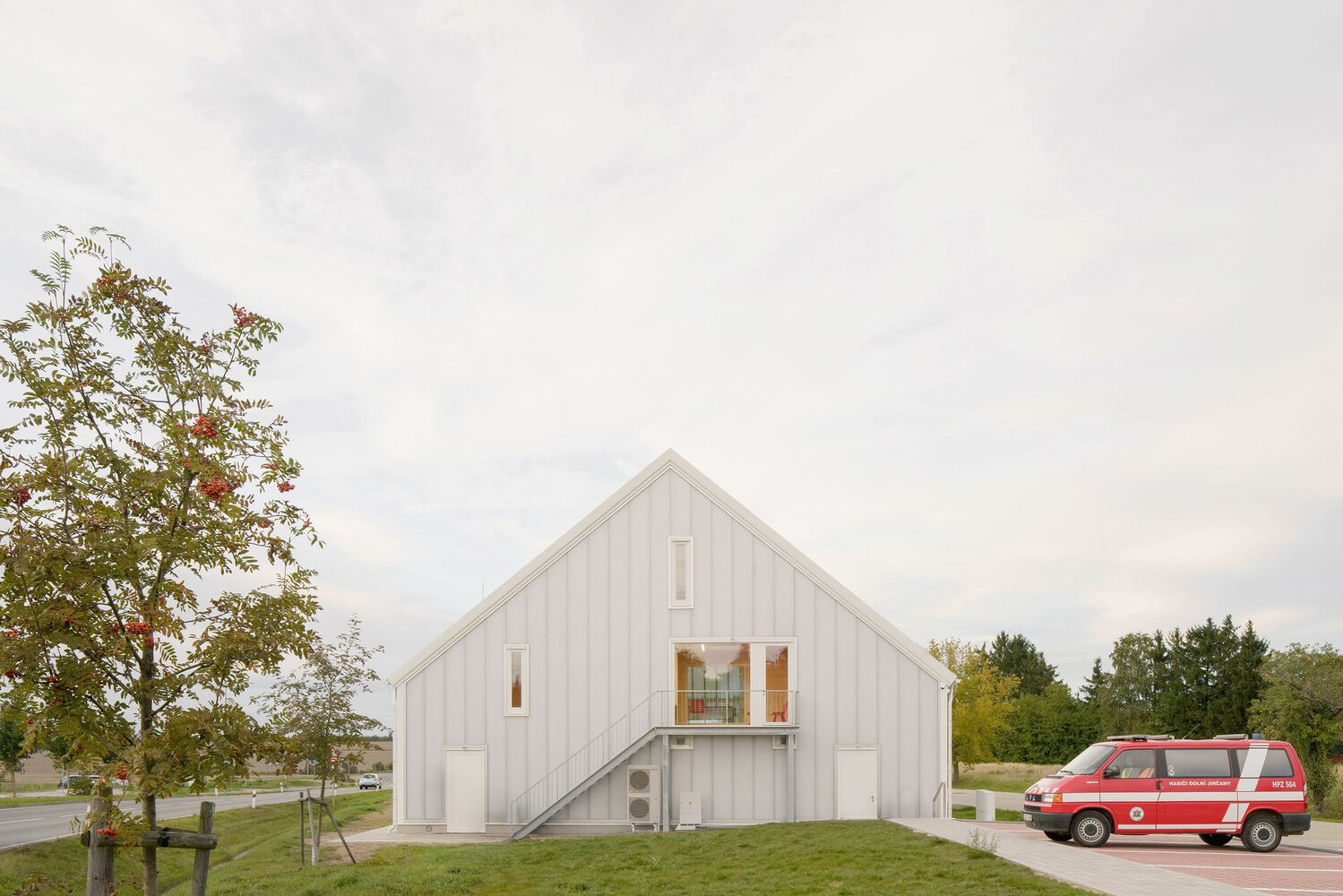White vertical-clad gabled structure with external staircase on grass slope beside parked emergency vehicle