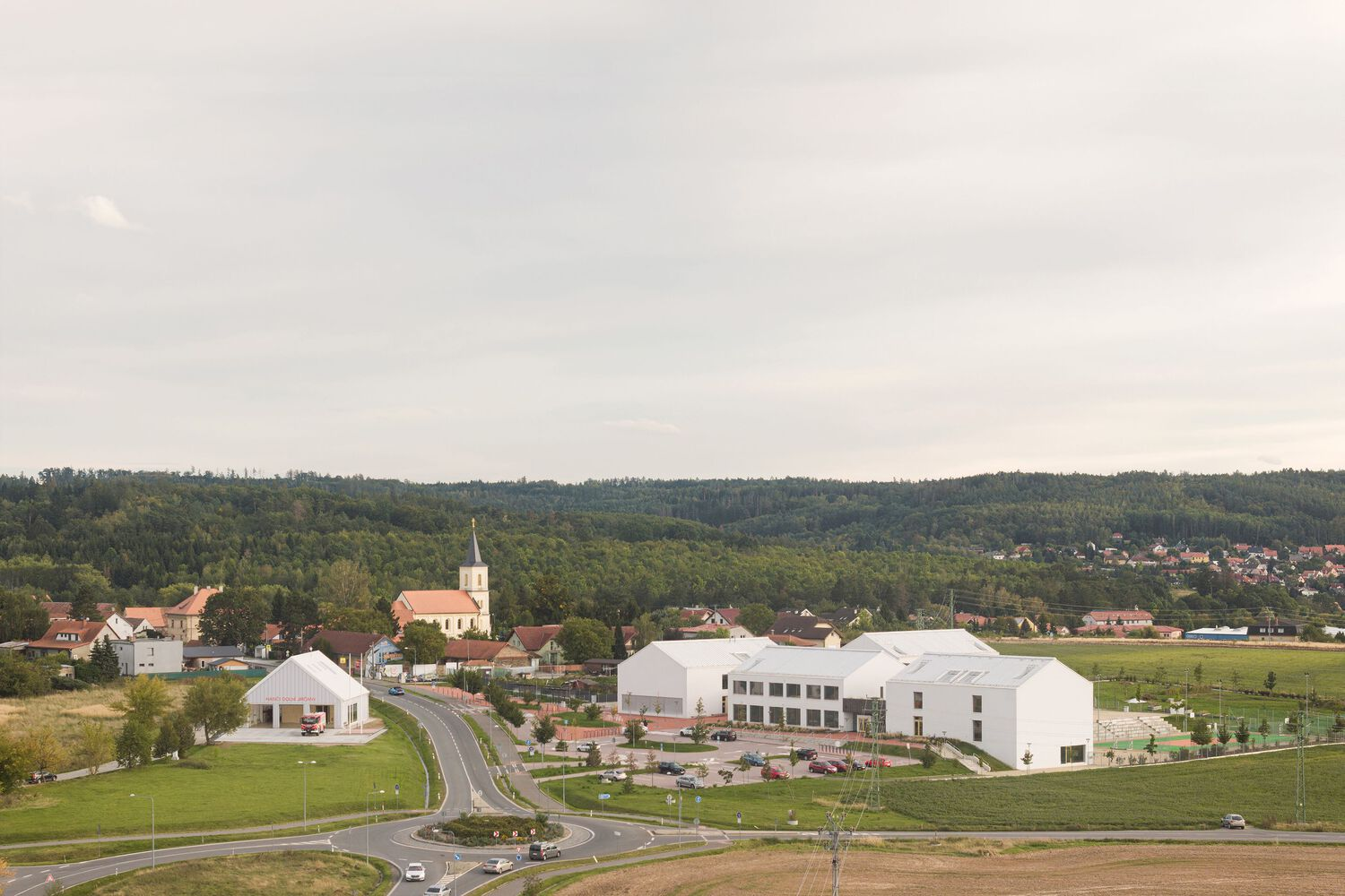 Aerial view of white gabled volumes set in a village landscape with forested hills beyond