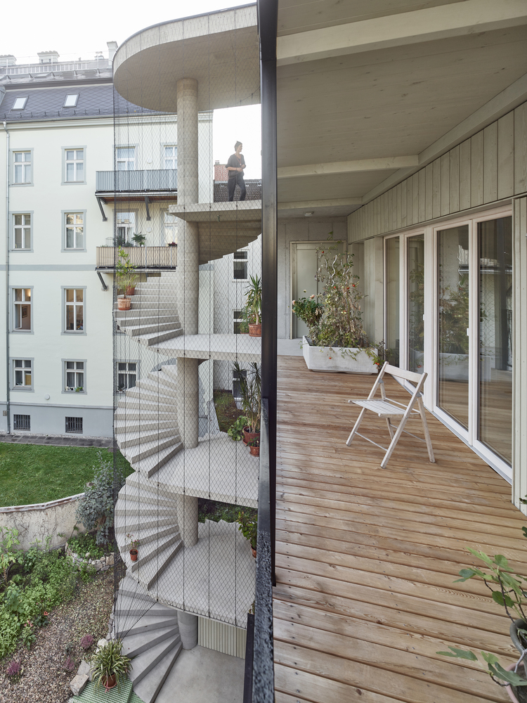 Timber balcony deck with white folding chair looking out toward the spiraling concrete stair tower clad in mesh