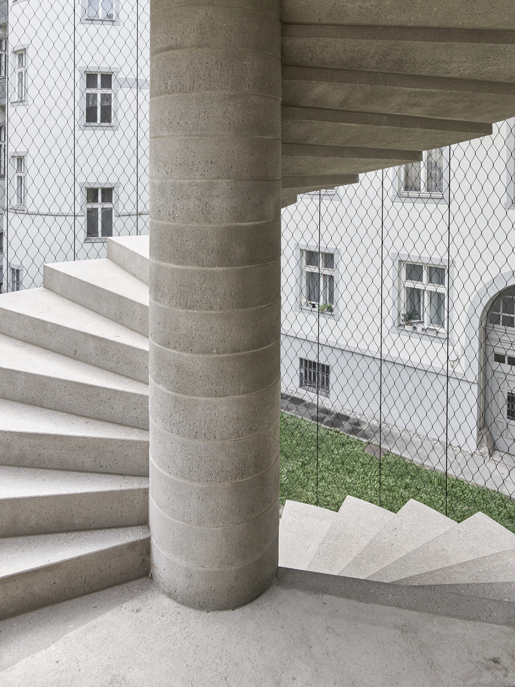 Spiral concrete stair wrapping a cylindrical column overlooking a mesh-screened courtyard with grass