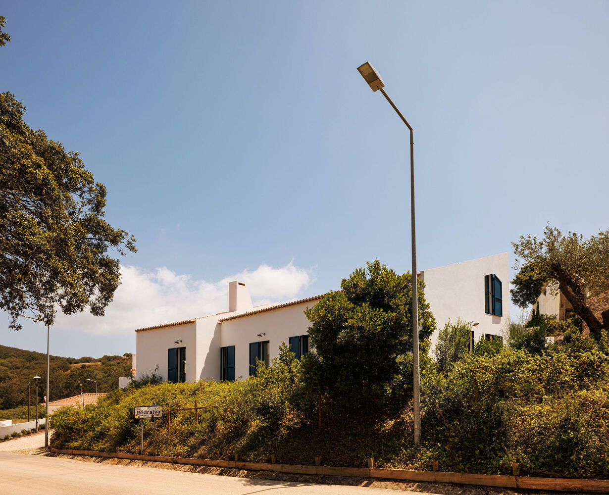 Street view showing white volumes with clay tile roof nestled among olive trees on sloping terrain
