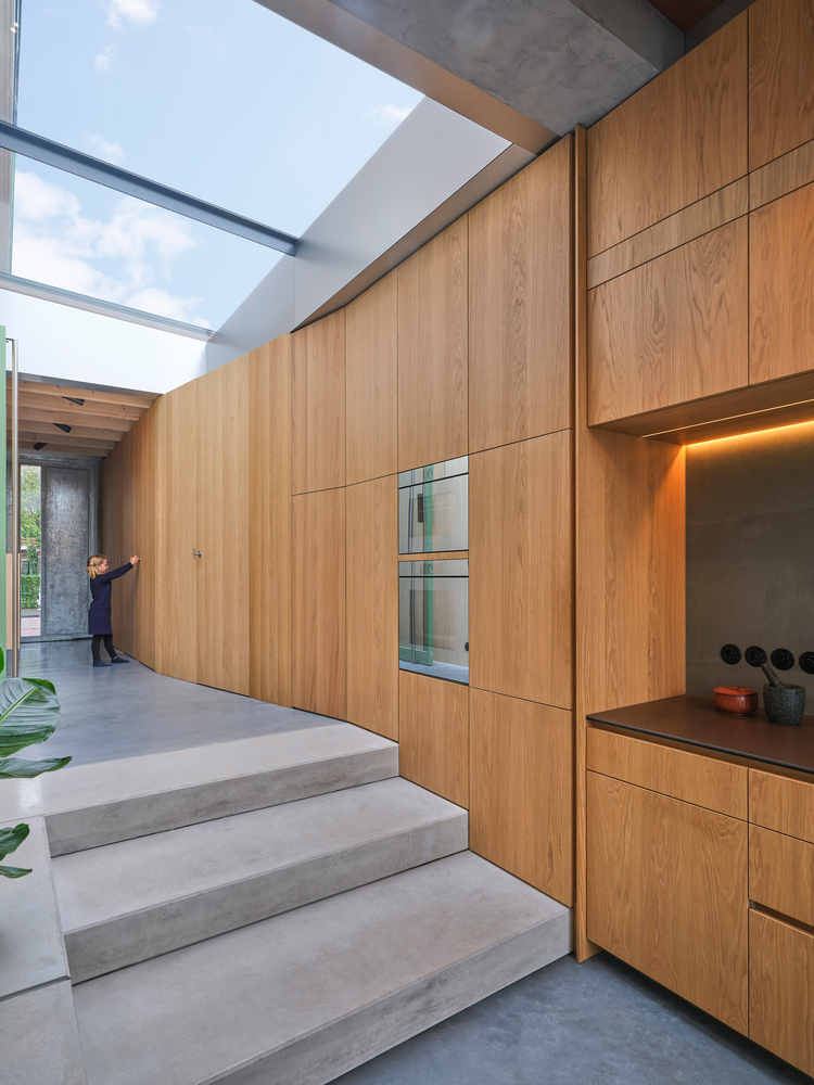 Concrete stairs rising alongside a full-height oak cabinetry wall beneath a skylight with a person in the distance