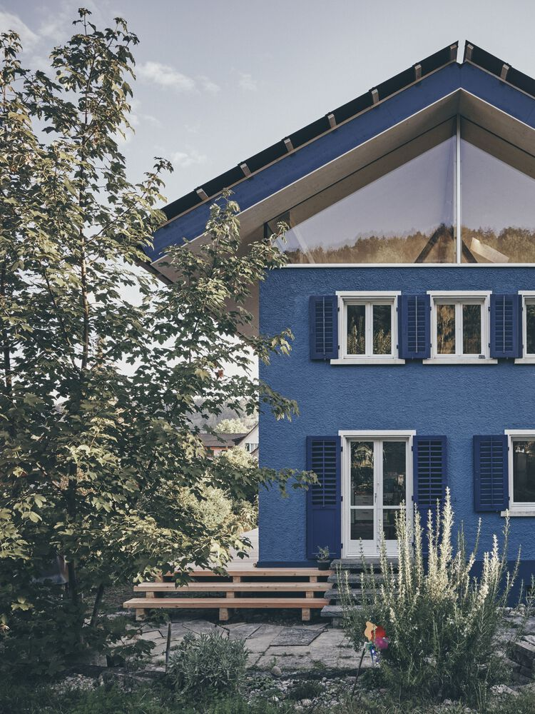 Garden facade with blue plaster walls, white-framed windows, dark shutters and timber bench beneath a young tree