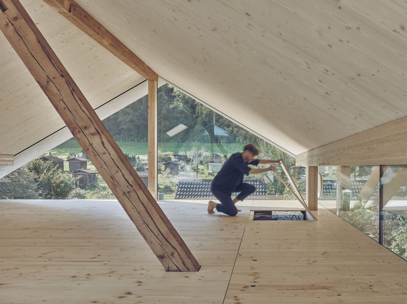 Attic room with plywood flooring, sloped timber beam, triangular window and figure working on the floor