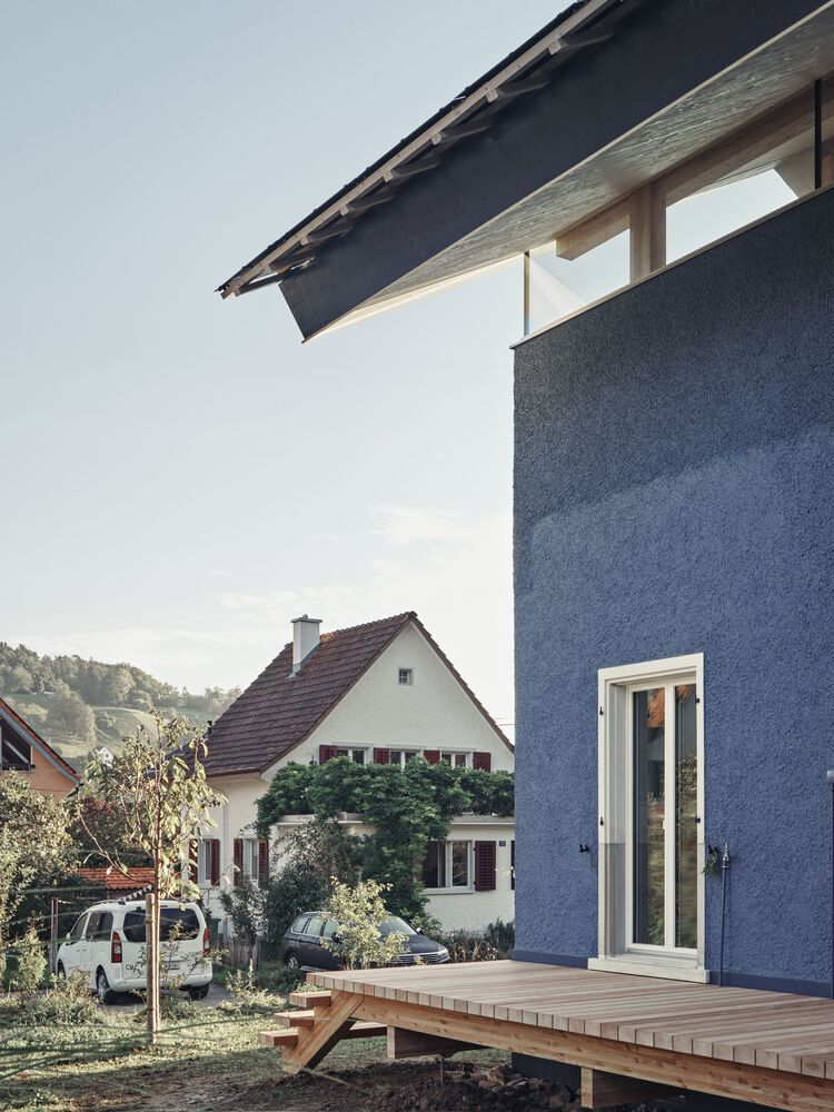 Blue rendered facade with timber deck and white-framed door overlooking neighboring gabled house