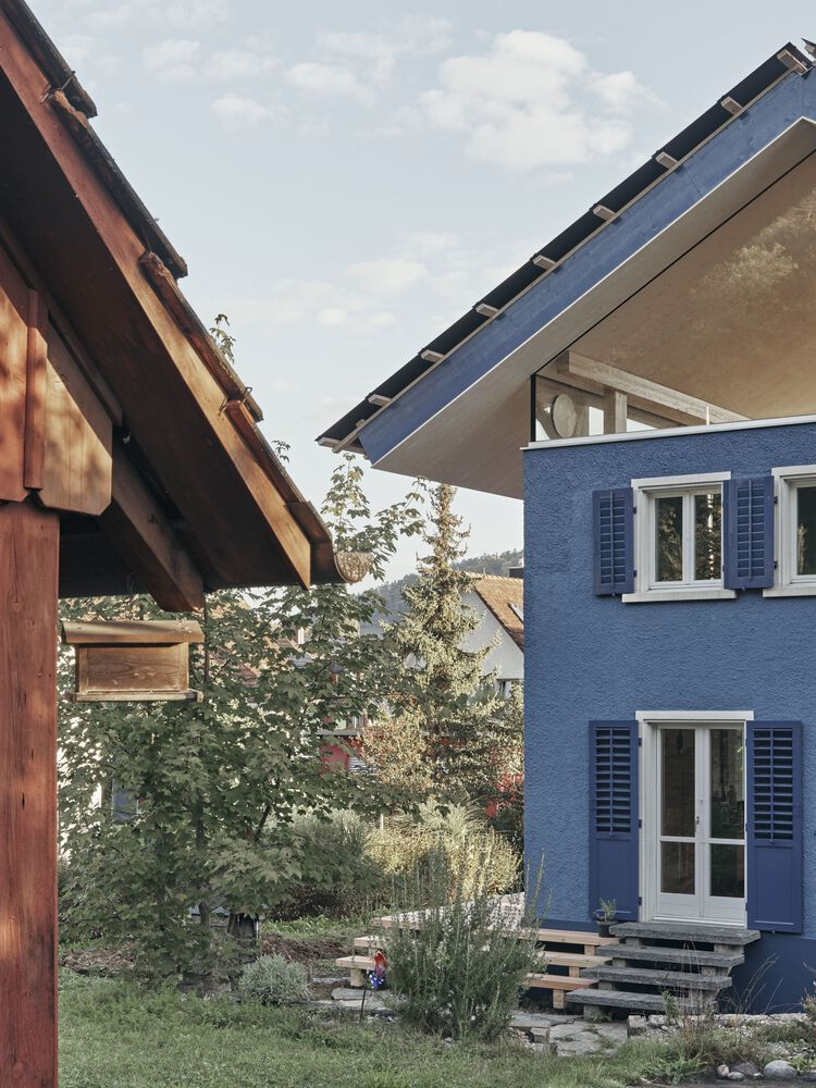 Blue stucco facade with white shuttered windows beside planted garden and timber structure
