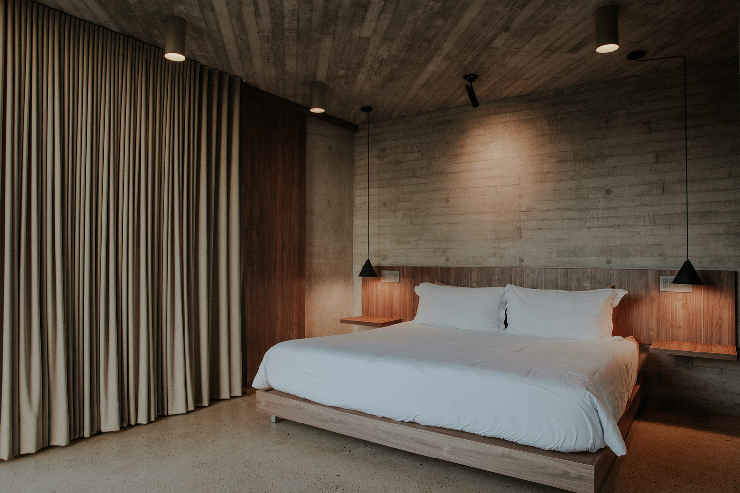 Bedroom with walnut headboard and nightstands beneath board-formed concrete walls and ceiling with pendant lights