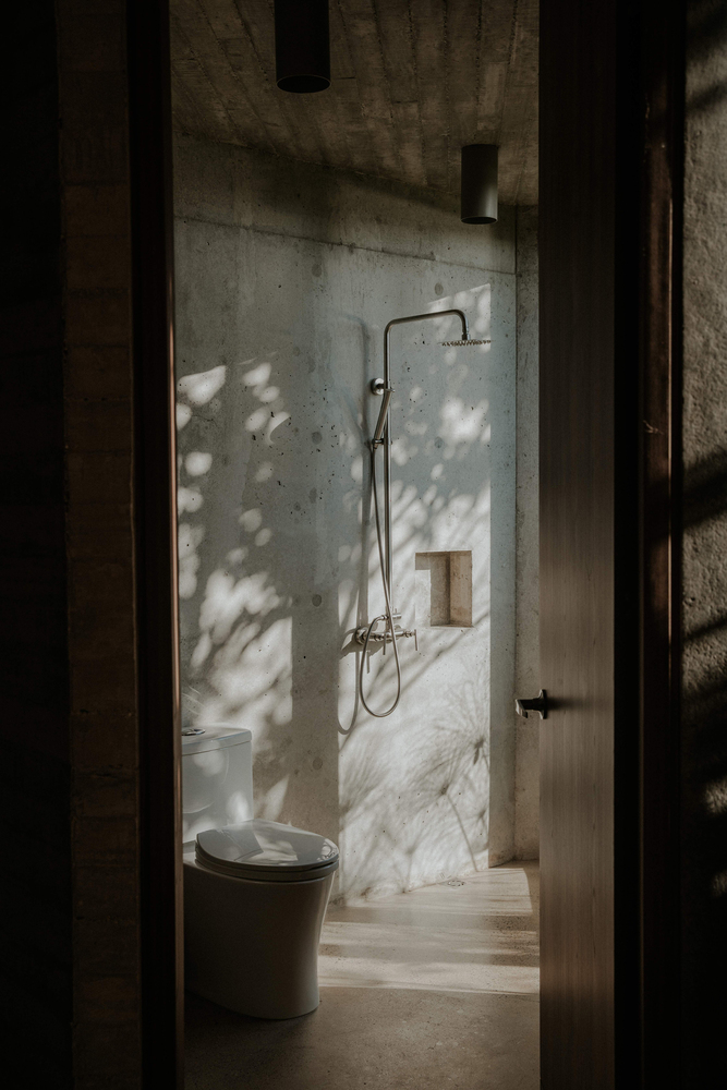 Bathroom doorway framing concrete and marble shower enclosure with dappled sunlight filtering through adjacent foliage