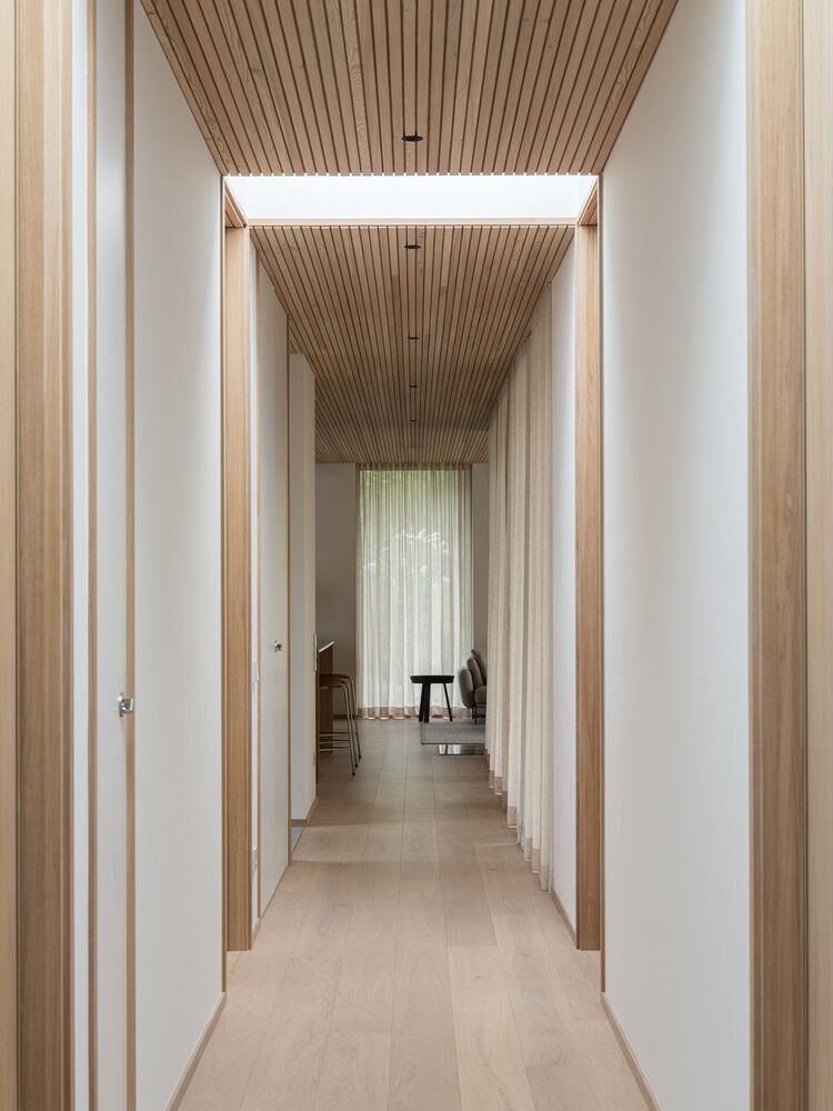 Long corridor with timber-slatted ceiling, recessed skylight, and white doors lining pale wood floors