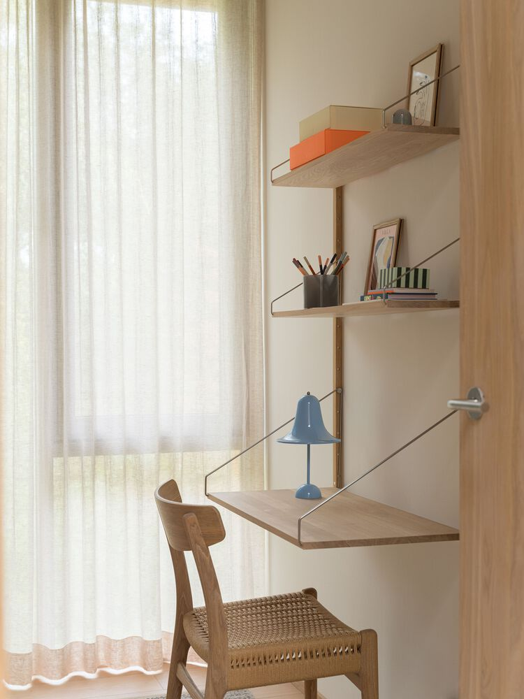 Wall-mounted desk shelves with wire rail brackets and cane chair beside a draped window