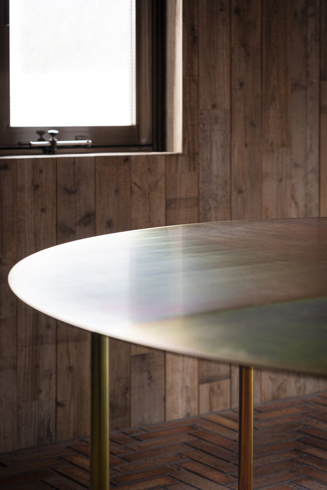 Oval marble tabletop on brass legs against vertical timber cladding with diffused daylight from a window