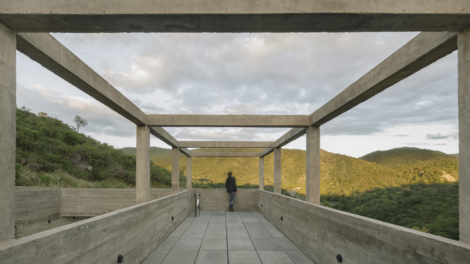 Open rooftop terrace framed by concrete columns with figure and dog looking toward sunlit hills