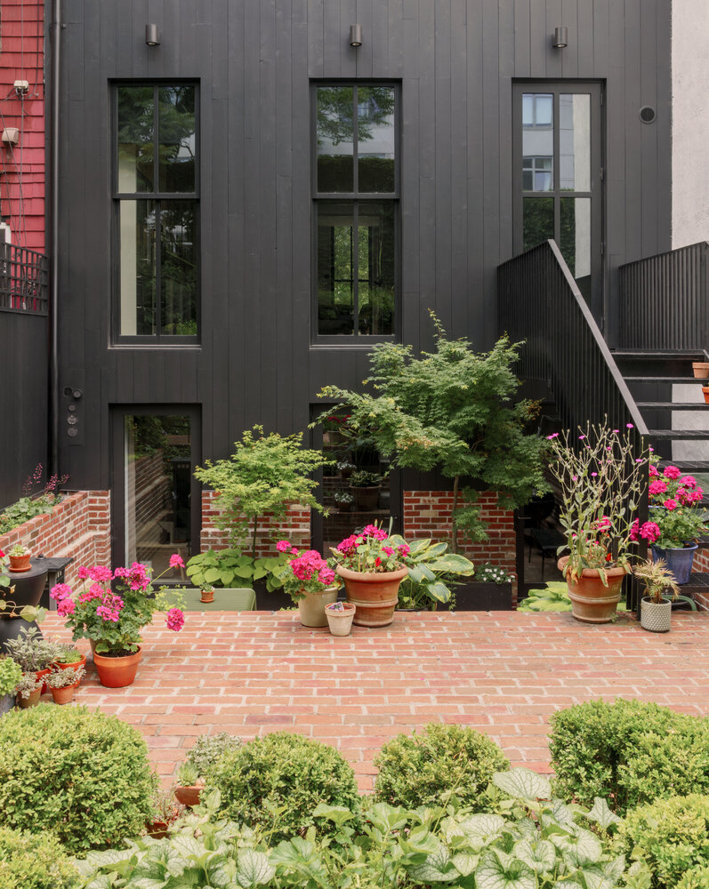 Dark vertical paneled facade with tall windows overlooking brick patio and potted flowering plants
