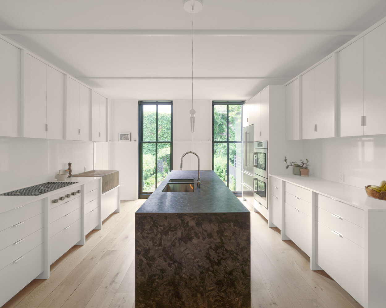 Galley kitchen with white cabinetry and dark stone island beneath twin windows facing greenery