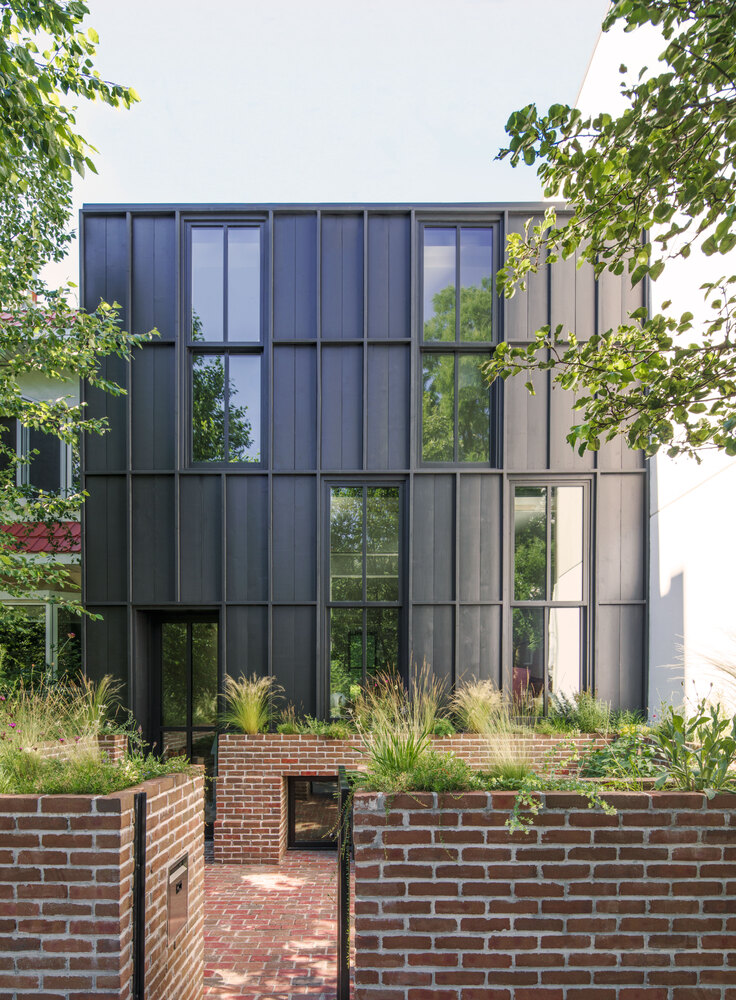 Street facade with black metal and glass panels rising above brick garden wall and grasses