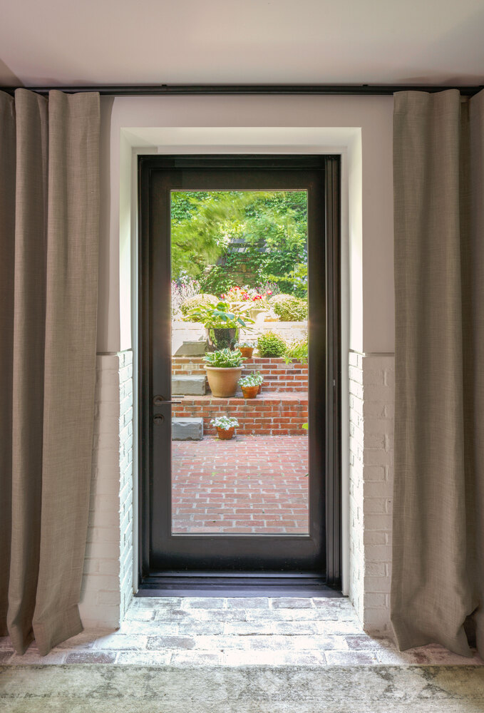Black-framed glass door with linen curtains opening onto a brick courtyard with potted plants