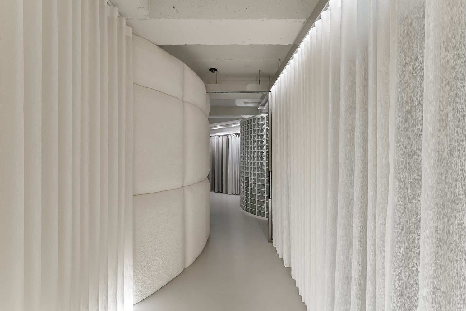 Narrow corridor defined by curved padded wall and white vertical curtains under concrete soffit