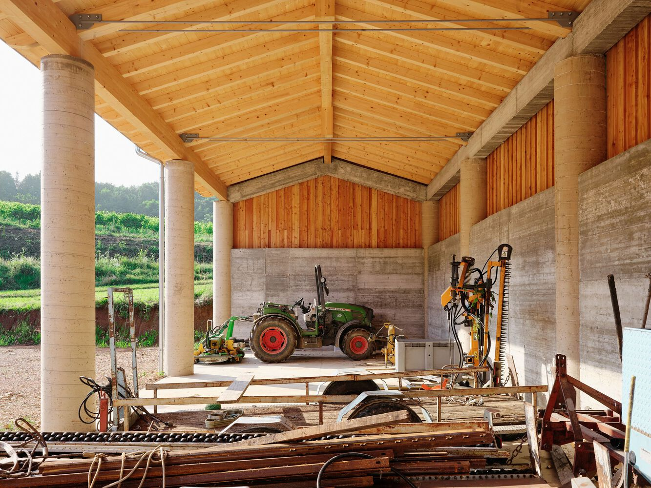 Open-sided storage bay with exposed timber beams and rammed earth walls sheltering a tractor and equipment