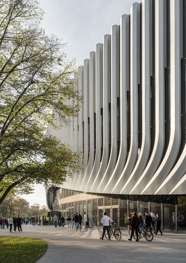 Ground-level view of the glass entrance beneath the sweeping white vertical fins with pedestrians and cyclists
