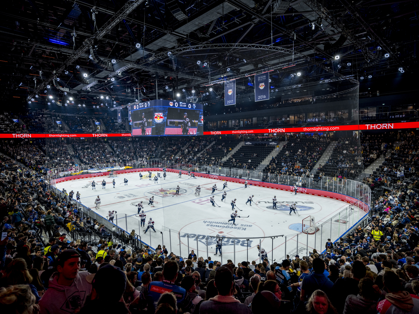 Wide view of the arena bowl with exposed steel roof trusses and red perimeter boards during a game
