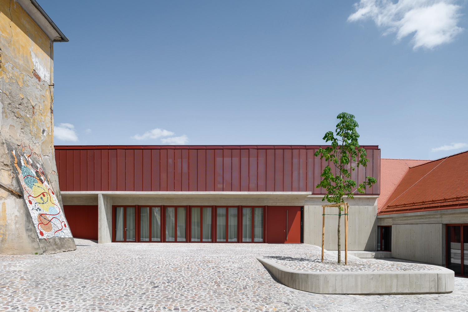 Courtyard view with red metal-clad facade and curved concrete planter holding a young tree
