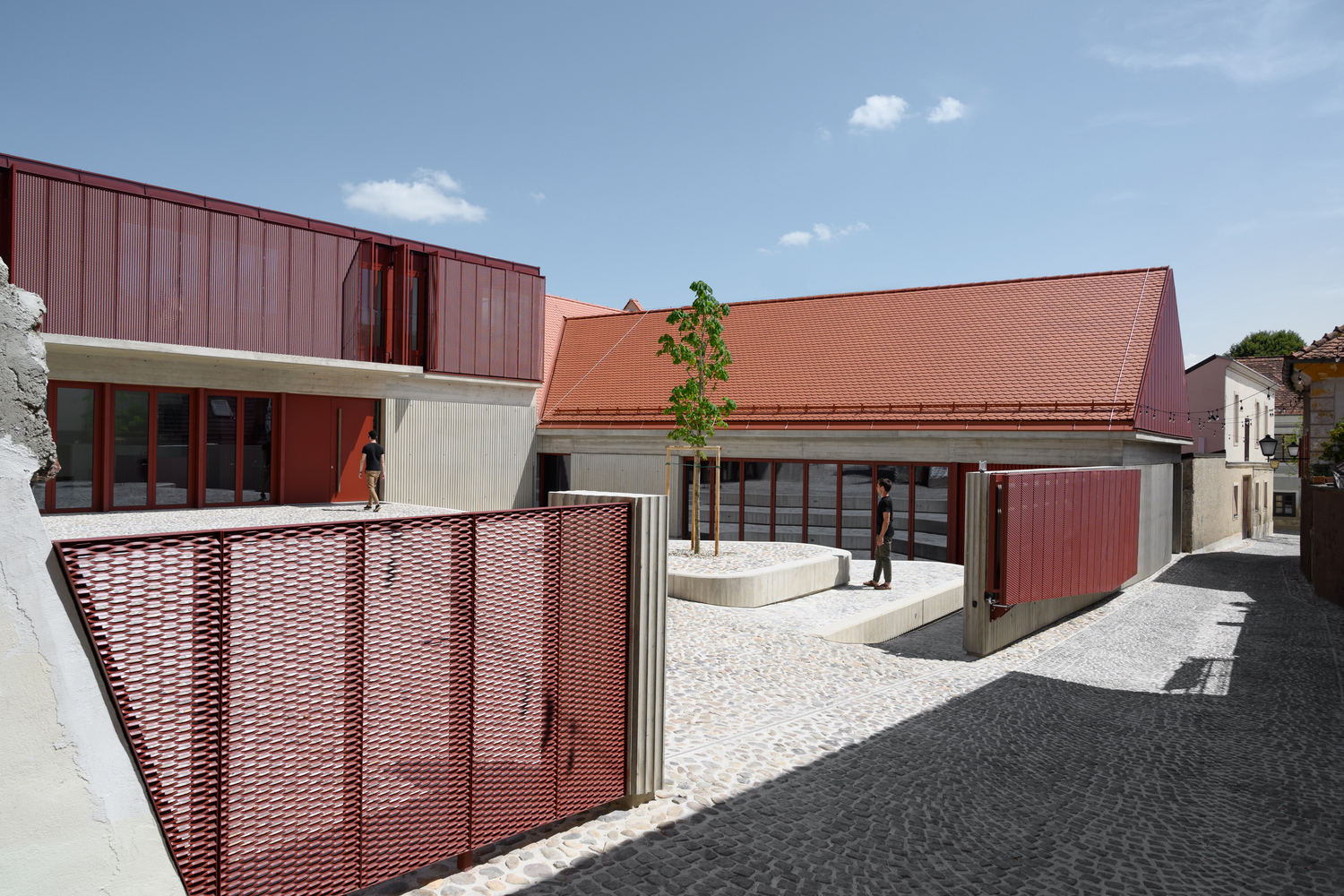 Cobblestone courtyard showing perforated red metal screen fence and sloped tile roof under blue sky