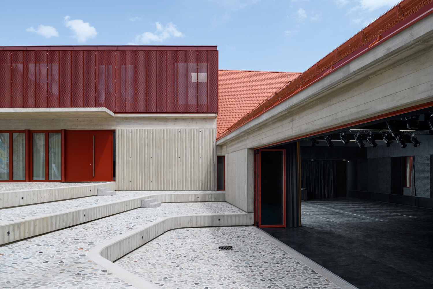 Courtyard with terrazzo paving and sunken concrete seating steps facing red metal cladding and orange tile roof