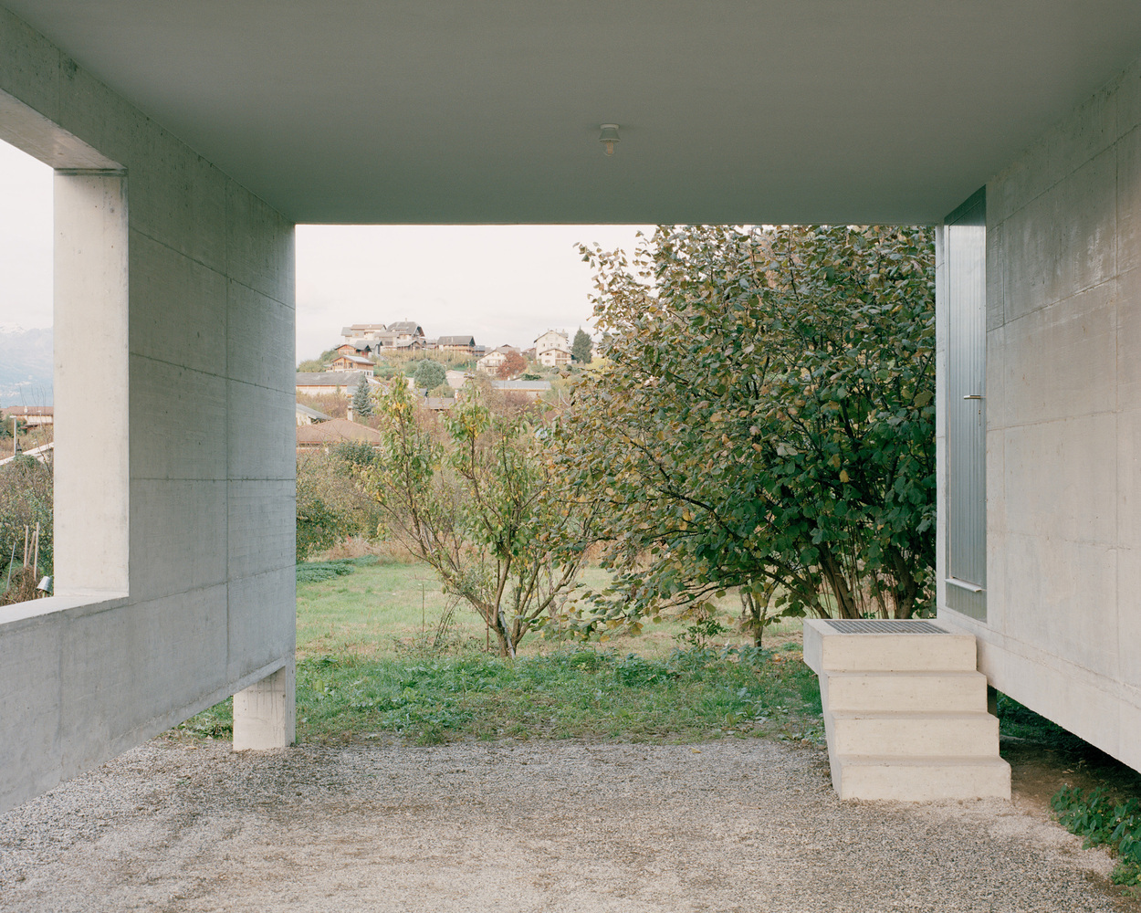 Covered entrance portico framed by concrete walls opening to fruit trees and a hillside village