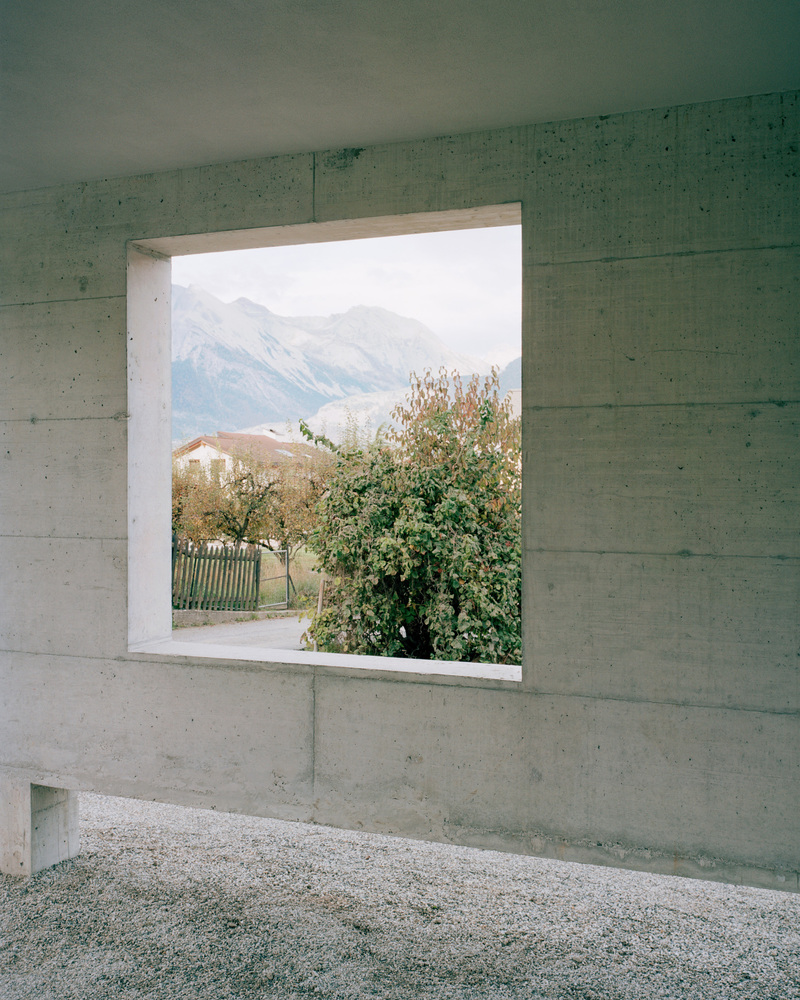 Square concrete window framing a view of fruit trees and distant mountains under overcast skies